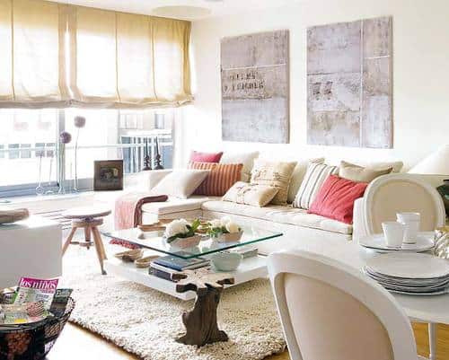 A neutral and bright living room with soft purple art and pink and neutral pillows and throw on a white sectional sofa.  A Glass coffee table and small dining table. 