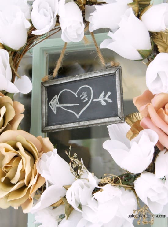 A cute Dollar Tree valentine wreath made with white roses and gold stems hang on a mint green french door with a little chalkboard sign in the middle.  