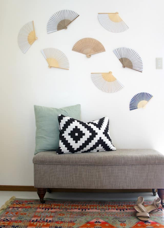 Boho fan wall art with dollar store folding fans hang on a white wall above a grey storage bench and colorful rug. 