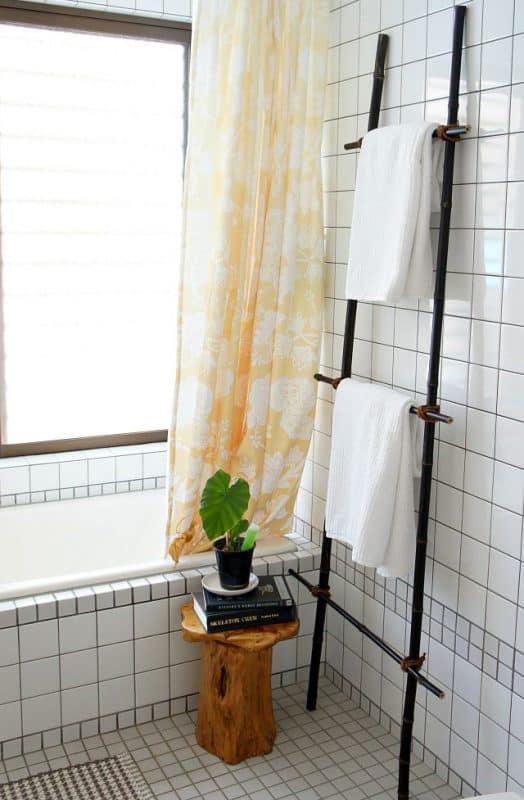 A DIY bamboo ladder holds towels in a white bathroom with a large window, yellow shower curtain, and wood sump table with plant. 