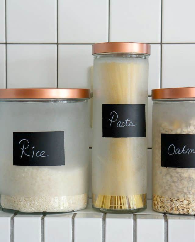 An ikea glass canister hack with copper lids and chalkboard paint labels.  Three canisters set on a tiled shelf with rice, pasta, and oatmeal. 