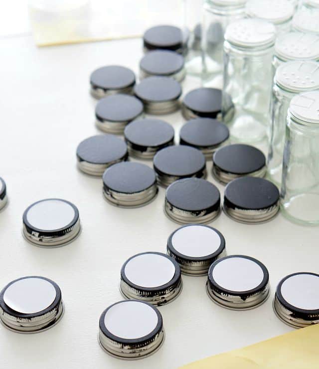 Chalkboard painted spice jar lids are on a white table with white labels on some of them. 