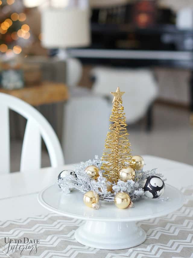 A small gold wire Christmas tree sits in small silver wreath with gold and black and white ornaments on a cake stand for a simple kitchen table centerpiece. 