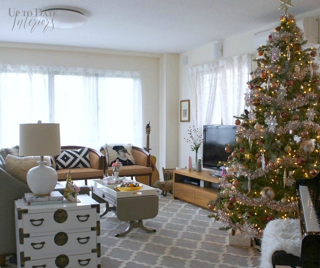 A wide view of a bright living room in Okinawa, Japan decorated for a holiday home tour showing a Christmas tree with tinsel garland and lots of pink and copper ornaments next to a piano and tv on modern wood media cabinet.  A large grey and white trellis rug anchor the seating area with two camel colored barrel chairs, grey sofa, and white campaign style side table.  Lots of windows with white sheer curtains keep the room light and airy. 