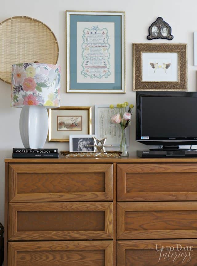 A walnut government issued dresser is decorated with art around a small table top tv, floral lamp, and decorating accessories. 
