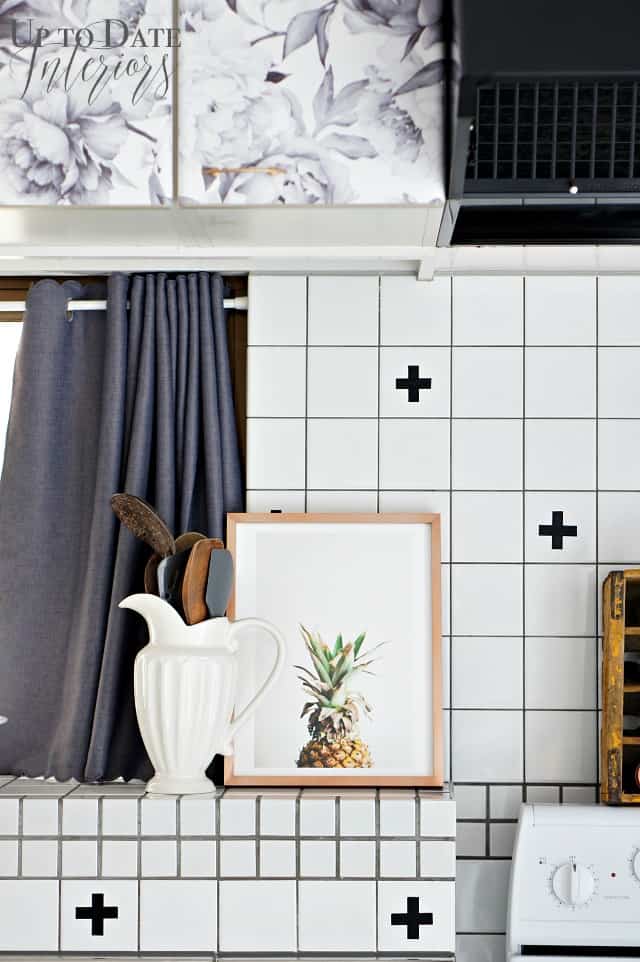 Minted art pineapple print is featured in a rental kitchen before and after makeover sitting on a white tile ledge with black swiss crosses on the tiles made with washi tape. A white ceramic pitcher holds utensils. 