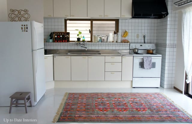 A Japanese rental home kitchen in Okinawa, Japan for Americans with white laminate floor, boho rug, white appliances and kitchen tile, metal counters, and cream cabinets.  The room has lots of natural light. 
