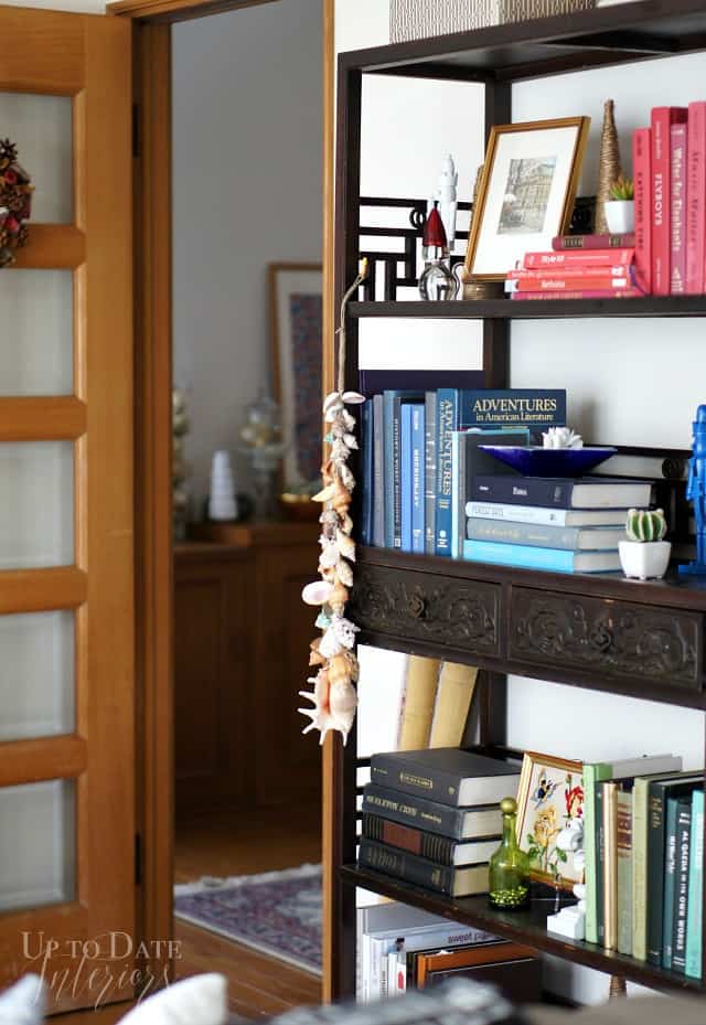 A colorfully decorated wood Asian eterege with red, blue, and green books and Christmas decor with a string of seashells hanging on the side is next to a pretty wood French door that leads into the Japanese foyer. 
