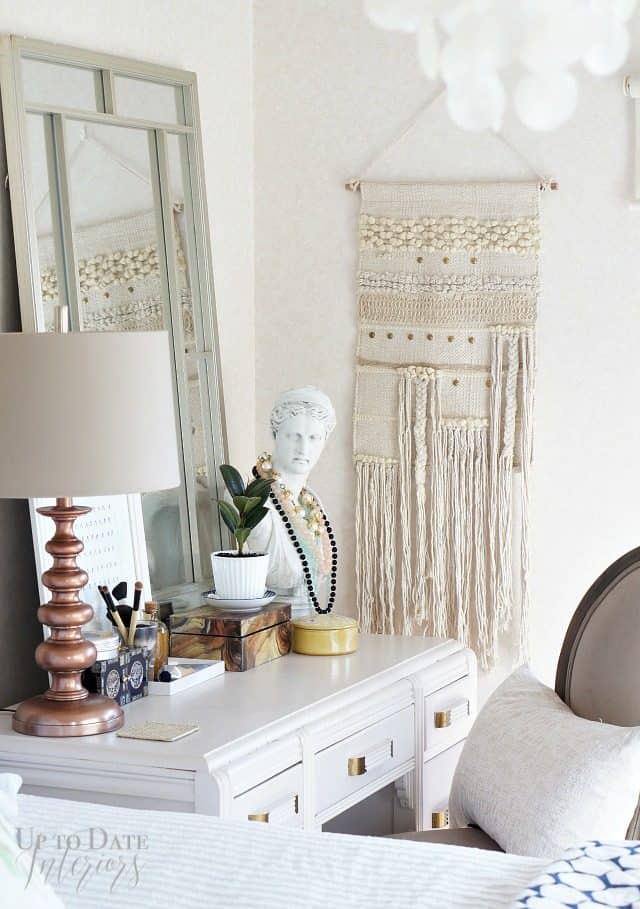 A renter friendly bedroom on a budget showing a cute neutral corner with a desk as a vanity with chair, pillow, and tall mirror.  A cute boho wall hanging in on the corner wall. 
