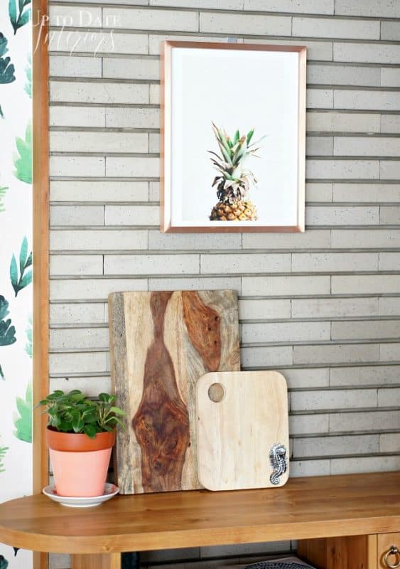A cute vignette in a Japanese kitchen showing a wood caddy storage cabinet with wood cutting boards leaning on a grey tile wall with a pineapple print hanging above in a copper frame.  A cute plant in a pink pot sits on the counter and a glimpse of botanic wallpaper on the side. 