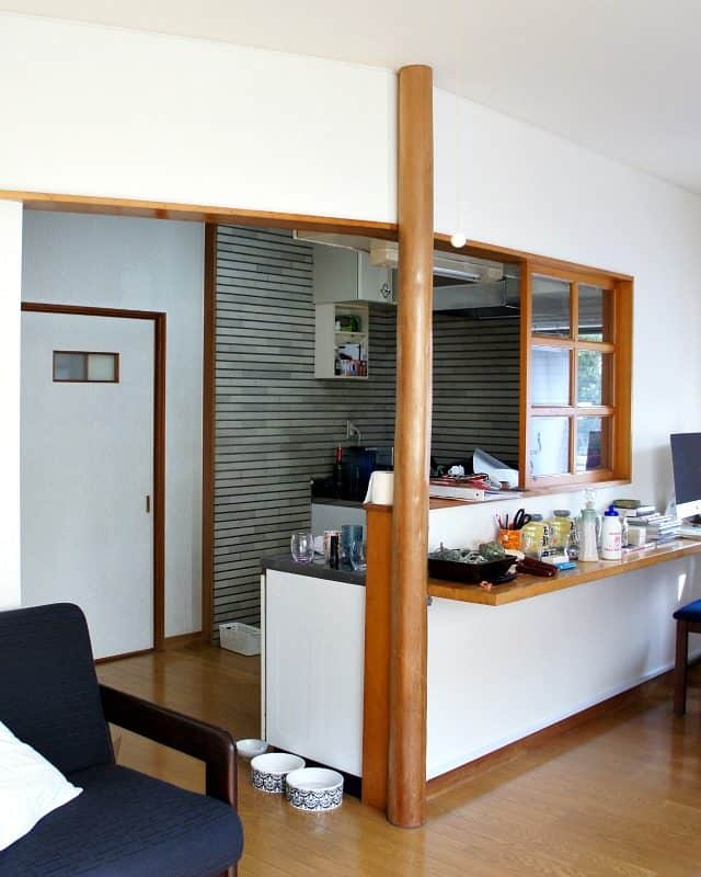 A rental kitchen before picture in Japan showing the opening into a small kitchen with grey tiled walls and white wallpaper sliding doors.  The opening has a cut out with a wood French window and a wood breakfast bar opening into the living room. 