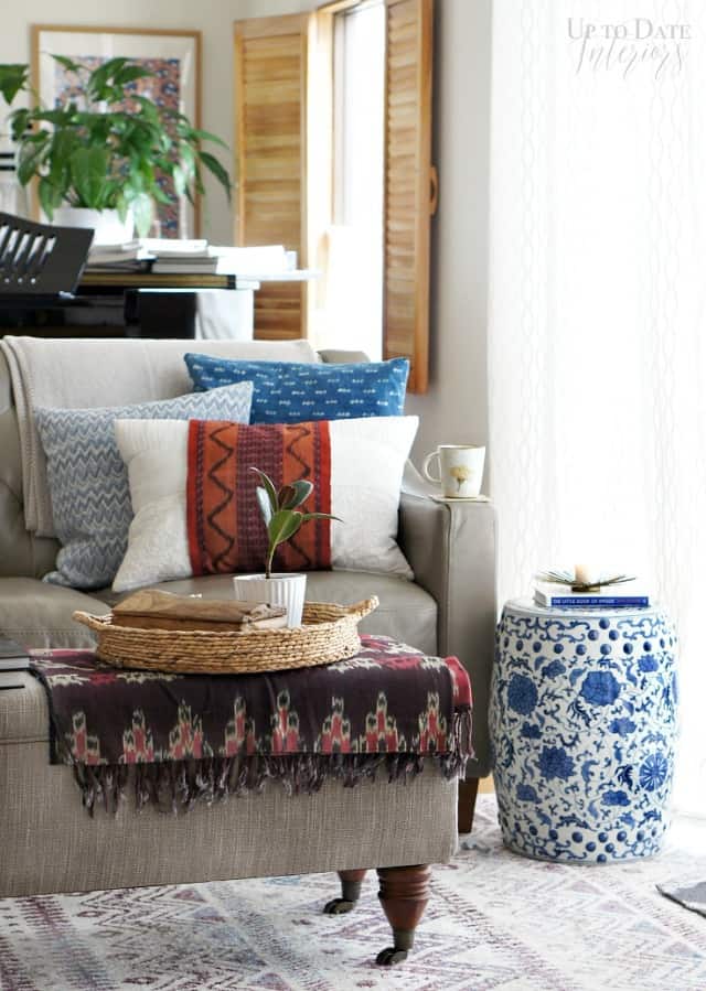 A colorful and eclectic living room view of a grey leather sofa in front of wood shutters and grand piano.  A chinoiserie garden stool is used as a sofa table and a grey upholstered storage bench with brass wheels is used as a coffee table.  Global pillows and textiles give it a hygge style. 