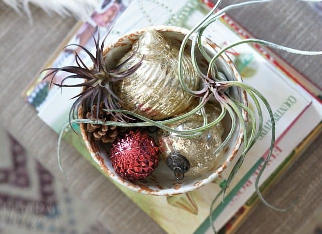 A birds eye view of a bowl of ornaments and airplants sitting on a stack of books on top of a grey upholstered bench. 