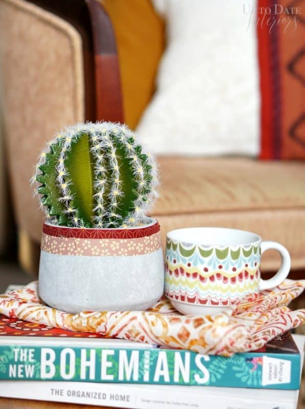A global concrete small planter with a faux cactus is on a stack of books with a colorful mug. 