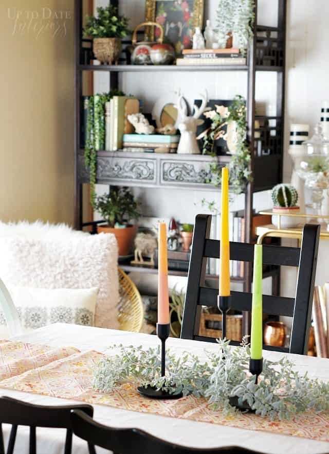Candles on a dining table with a boho eclectic bookcase in the corner with lots of books, plants, and Christmas decor. 