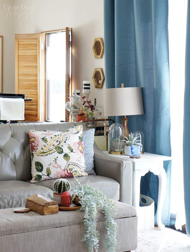 An eclectic and cozy living room with hidden remote storage on top of a storage bench ottoman, blue curtains, and wooden window shutters. 