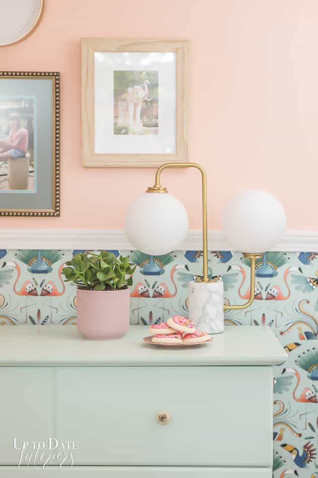 A mint dresser with a modern lamp, small plant, and cookies are in front of bird wallpaper. 