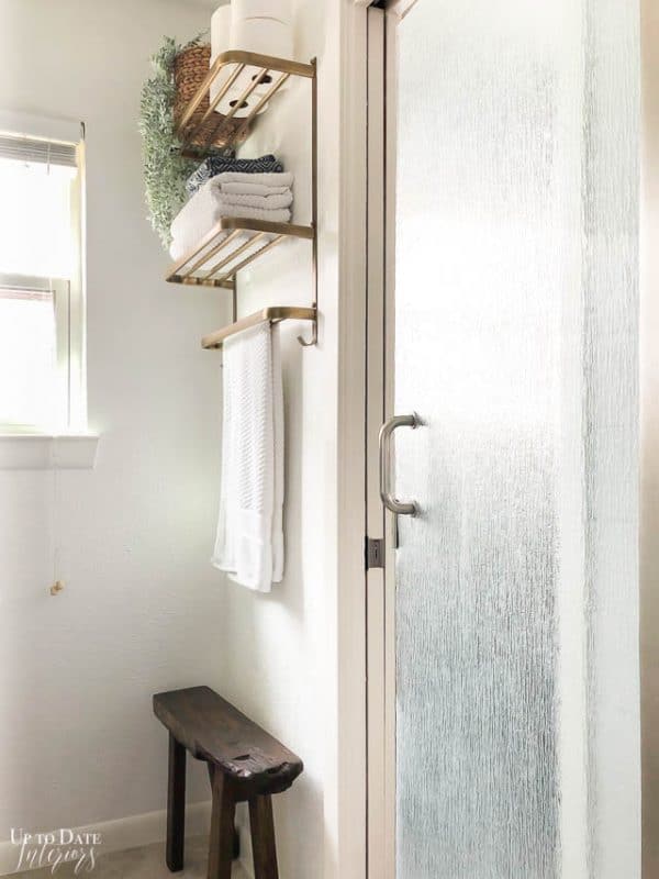 A small bathroom makeover showing white walls with a gold hotel style towel rack and small wood rustic bench. 