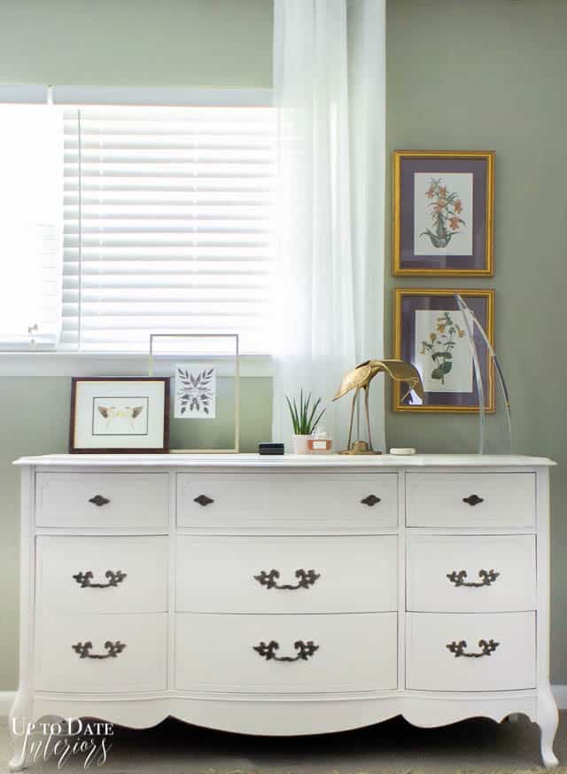 How to paint a dresser white!  White French provincial style dresser against a window and green khaki walls with botanical art prints and decor styled on top of dresser including a brass crane and lucite sculpture. 