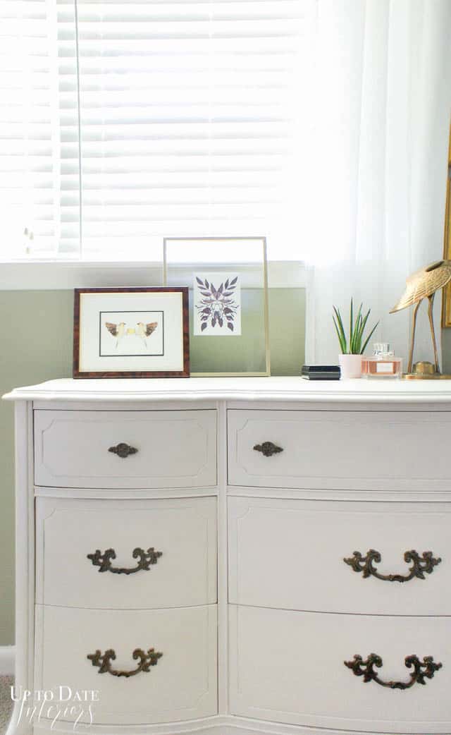 A close up picture of a dresser vignette in front of a window with a white dresser and khaki green walls and white sheer curtains and blinds. 