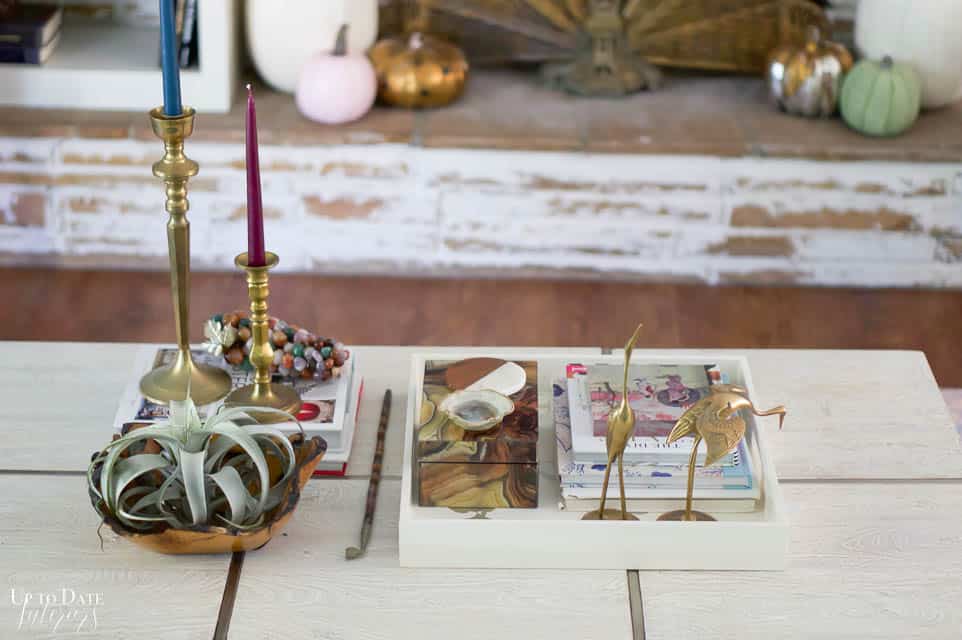 A close up look at a modern white and chrome coffee table with a large white tray holding brass cranes and books and a wood bowl with air plant, brass candlesticks with purple and navy taper candles, and decorative grapes sit on interior design books. 