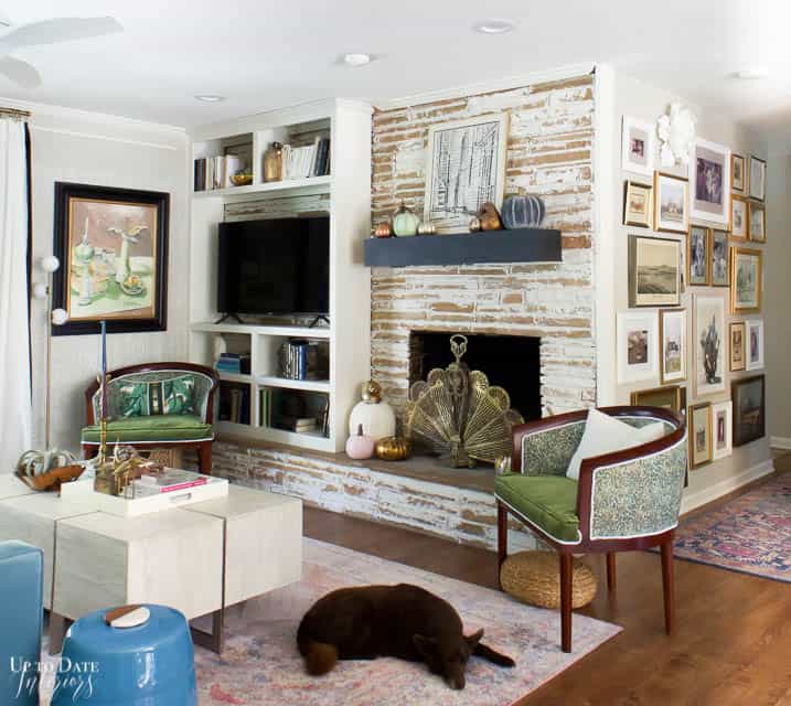 A full view of fireplace with german smear technique on tan stone and a built in bookcase for tv and decor.  A floating mantel in dark blue with pumpkins on top and black and white art.  You can see a full gallery wall and two green velvet chairs.  A large white coffee table with more Fall decor sits on a pink rug. 