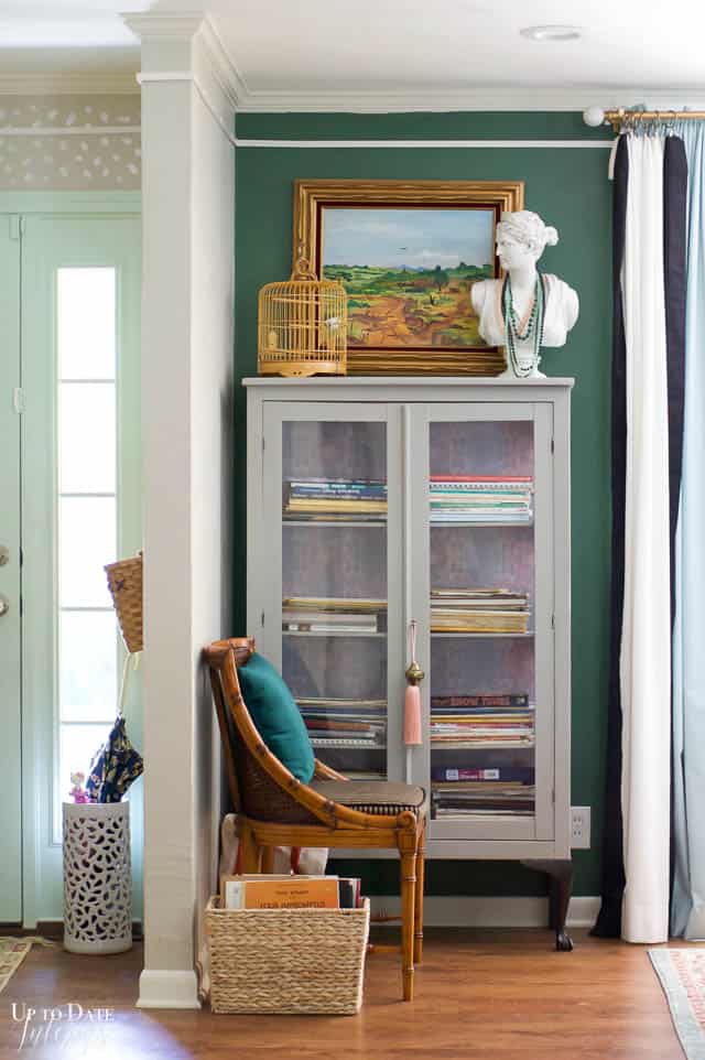 A grey display cabinet full of music books sits in front of a dark green wall with a beautiful oil landscape, bamboo birdcage, and woman's bust with colorful necklaces.  A pink tassel hangs on the cabinet lock and key.  There's a bamboo cane chair in front against the wall and a glimpse of the mint green front door sidelight.