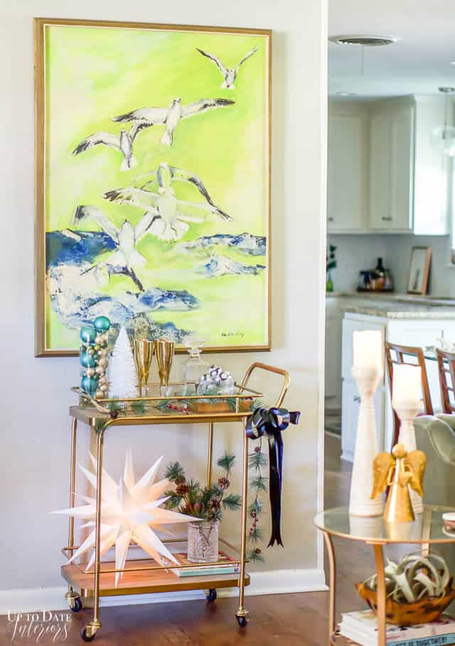 A large painting of birds in bright green is hanging above a gold bar cart decorated for Christmas. 
