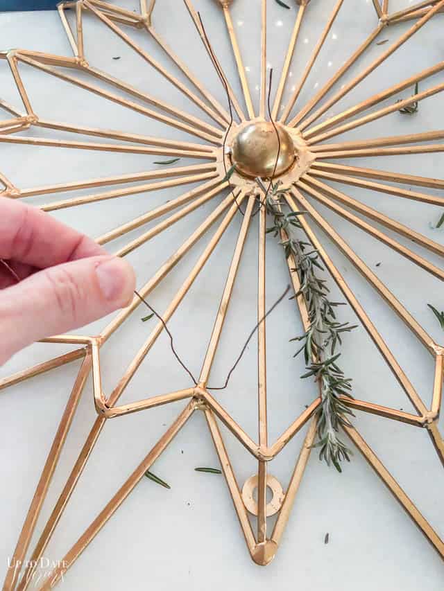 A beautiful gold star shaped wall hanging and a woman's hand wrapping florist wire to secure fresh rosemary to it to make a rosemary wreath.