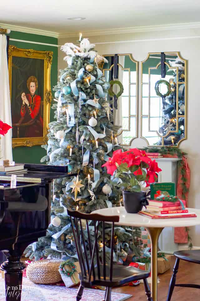 A beautiful blue ribbon and elegant Christmas ornaments decorated tree is in a corner with mirrors and woman's portrait on the wall.  You can see part of a grand piano and a small marble table with black farmhouse chairs. 