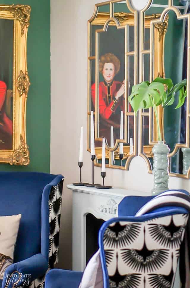 A close up view of a living room corner with jewel tones showing a pair of gold painted mirrors over a light blue mantel with two blue velvet wing back chairs and a green wall with a woman's portrait. 
