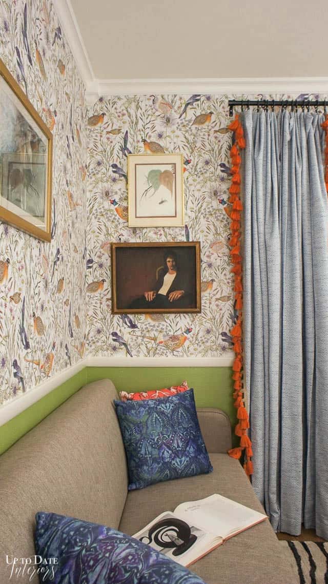 An office with bird wallpaper and curtains with trim, grey sofa with blue velvet pillows, and art on the walls. 