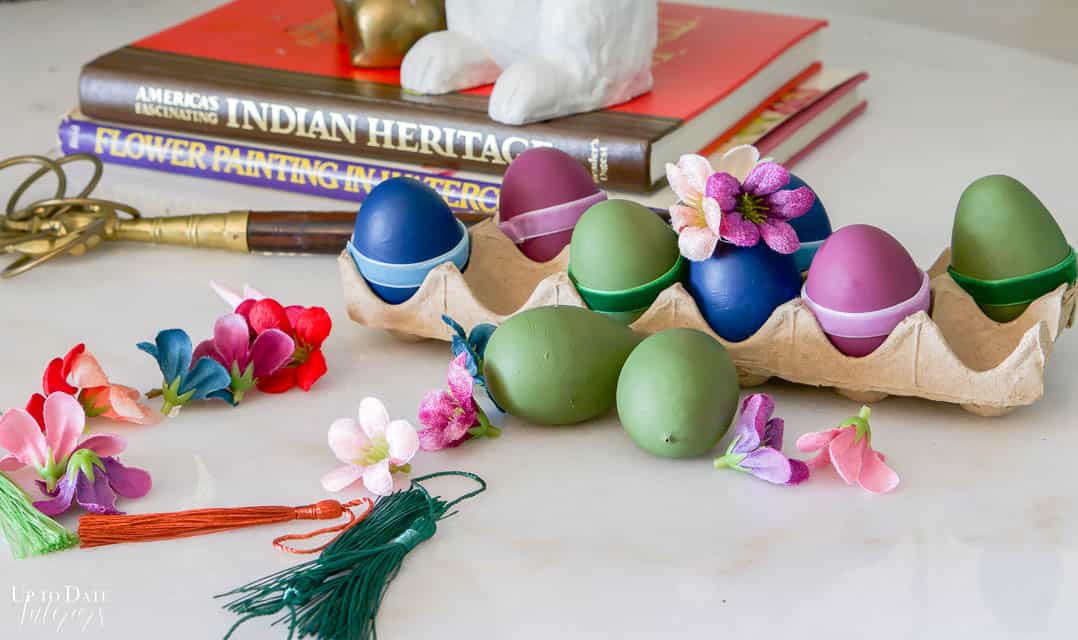 Decorate plastic eggs by painting them and hiding the seam with velvet ribbon.  A collection of eggs are displayed in a brown egg carton on a white marble table with small silk flowers and tassels. 