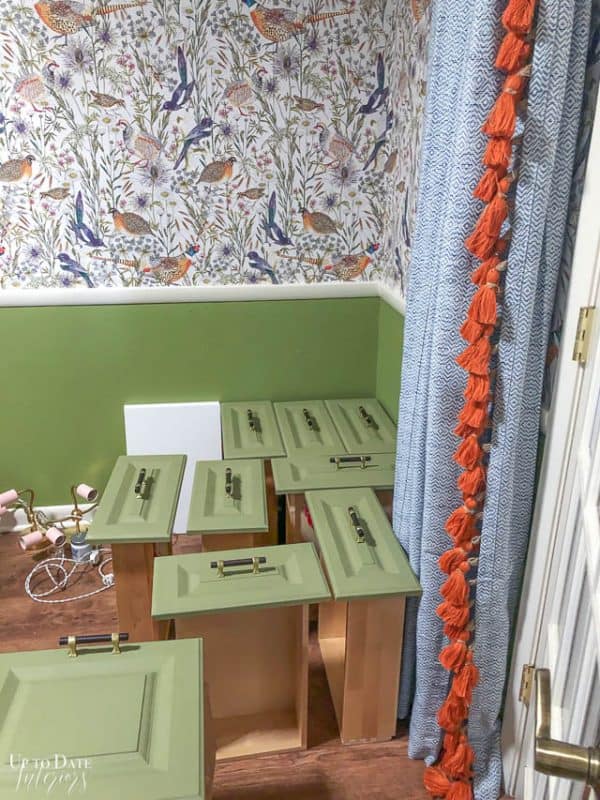 The best paint for office cabinets without sanding or priming in a pretty green color with wood floors, blue curtain with orange tassel, green wall paint below a white dado rail and bird wallpaper above a dado railing. 