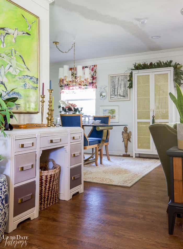 Green garland is beautifully draped over a white cane cabinet in a small dining area with bright pink floral roman shade and dark blue dining chairs on a pink rug.  View of art decor desk with bold green and blue art sit in the foreground. 