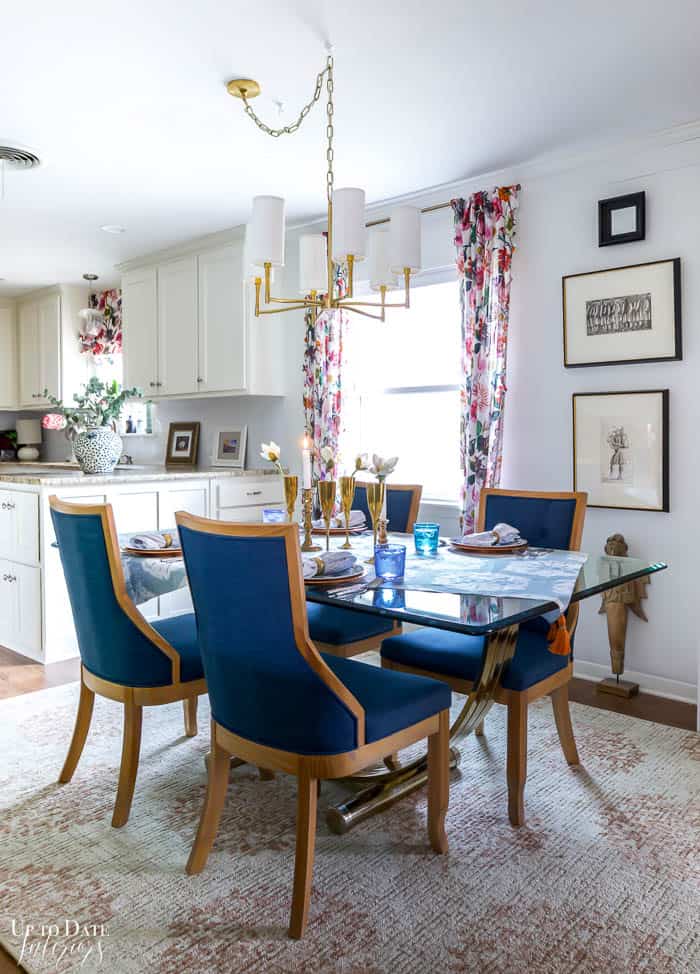 I added a cottagecore home decor to our dining room with colorful floral curtains on white walls with fine art.  A pink dining room rug anchors a glass and gold base table with vibrant blue upholstered wood country style chairs.  A gold and white chandelier completes the space and the table is all ready for a Spring meal. 
