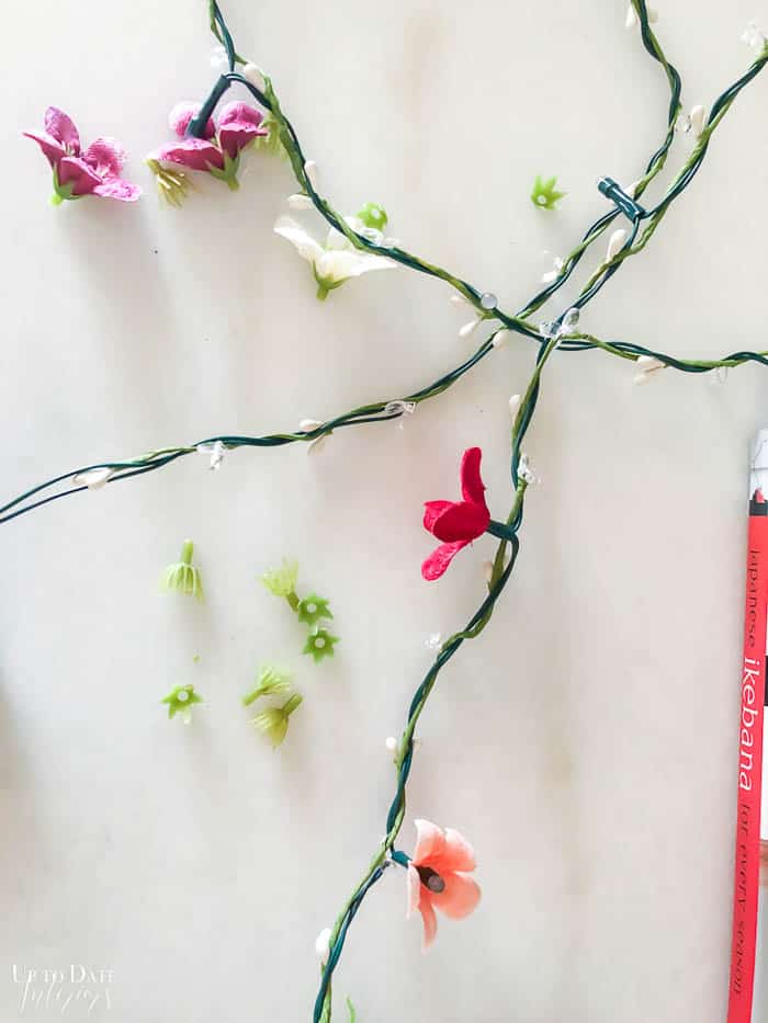 DIY flower fairy light garland step by step picture showing silk flower heads and how to remove the middle to attach to fairy string light bulb.  The string lights with flowers are on a white marble table. 