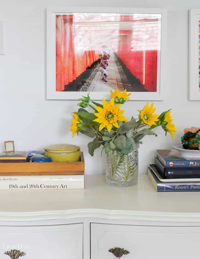 Fake sunflower stems in a crystal vase are on a white dresser with books and art. 