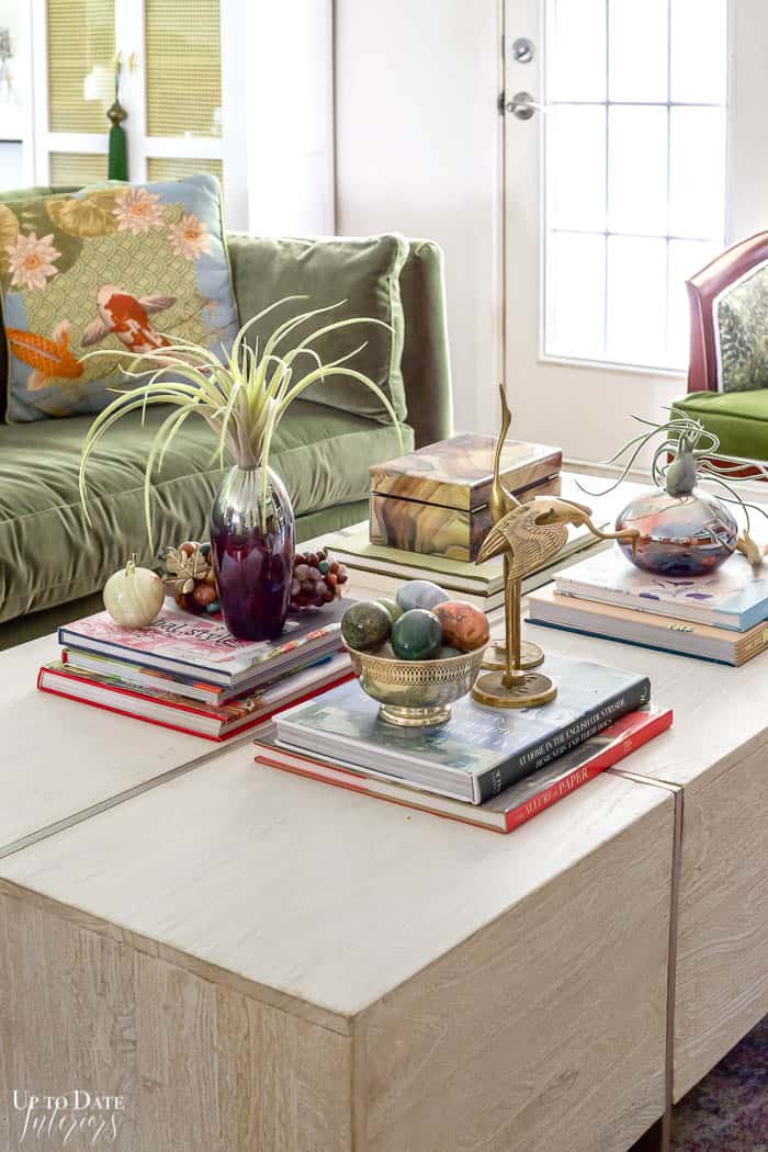 Colorful and eclectic stacked coffee table books styled on a large white rectangle coffee table with air plants, brass cranes, and colorful accessories. 