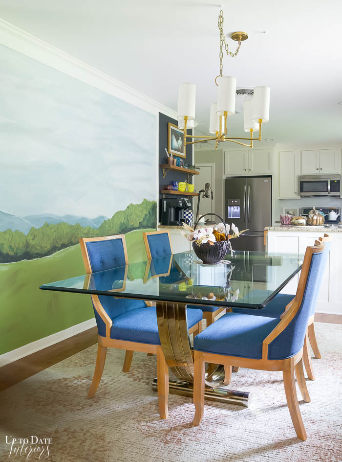 Small Dining area with glass top dining table and blue chairs and a landscape mural on the wall. View is into a white kitchen. 