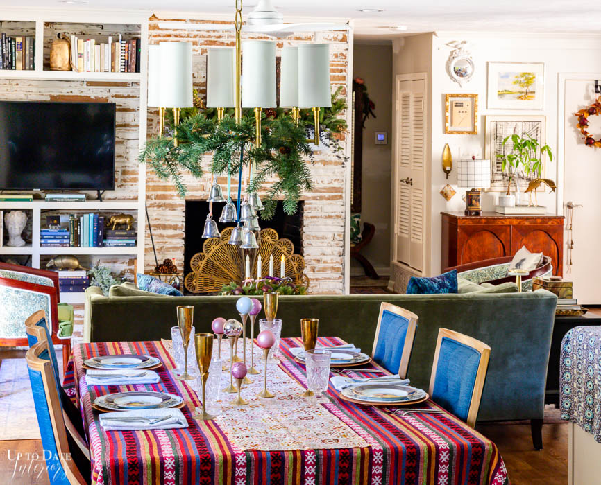 Mixed metals make a dreamy maximalist Christmas table setting with silver bells and greenery on a gold chandelier and brass champagne flutes and candlesticks with ornaments on a colorful tablecloth with blue chairs.  View of the open concept living areas is seen in the background. 