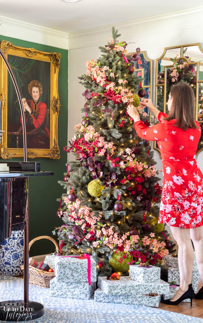 A picture demonstrating how to decorate a Christmas tree with flowers showing a woman in a red floral dress placing pink and red flowers in a spiral shape on a large tree that is fully decorated with lights, moss balls, and fruit ornaments.  Wrapped presents in botanical wrapping paper are under the tree.  You can see a woman's portrait on a green wall behind the tree in this maximalist living room setting. 
