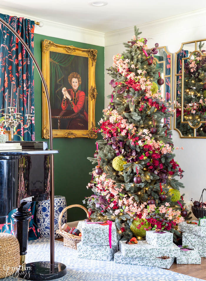A gorgeous colorful floral Christmas tree in a corner with presents wrapped in botanical wrapping paper and  woman's portrait on a dark green wall in the background. 