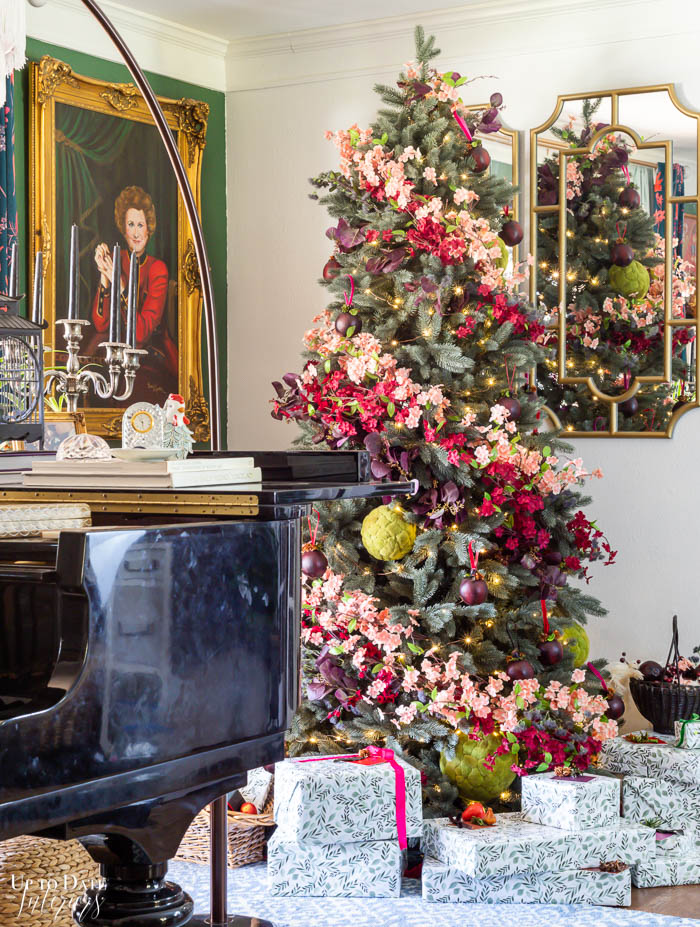 A baby grand piano sits near a floral decorated Christmas tree with a dark green accent wall and white wall with gold mirrors.  Wrapped presents are on the floor. 