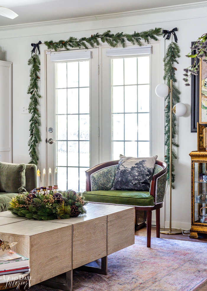 Pine garland hangs around white french doors and white walls next to gold furniture and a green velvet sofa and chair. 