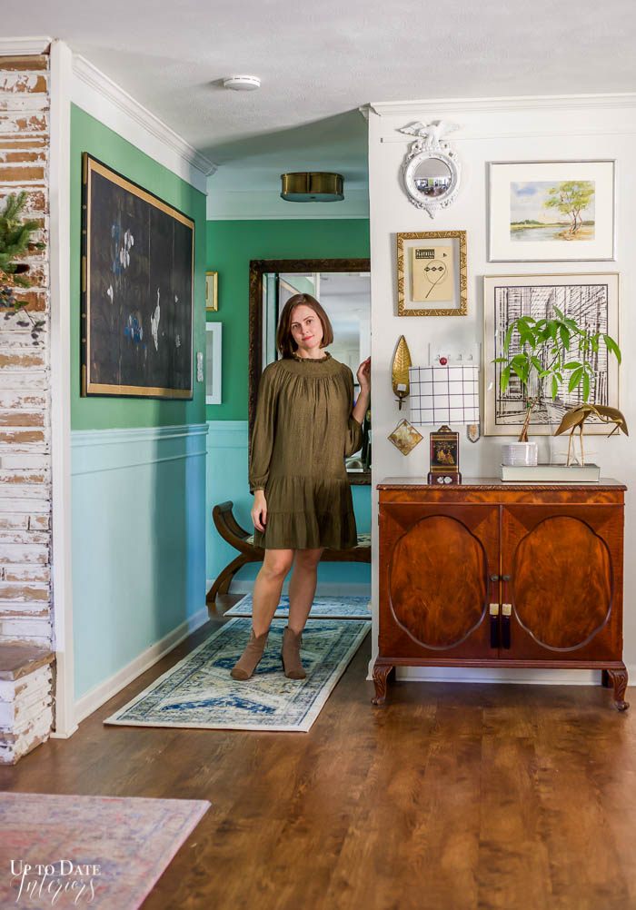 A woman standing in a blue and green hallway next to a bright white foyer with lots of art and maximalist style. 