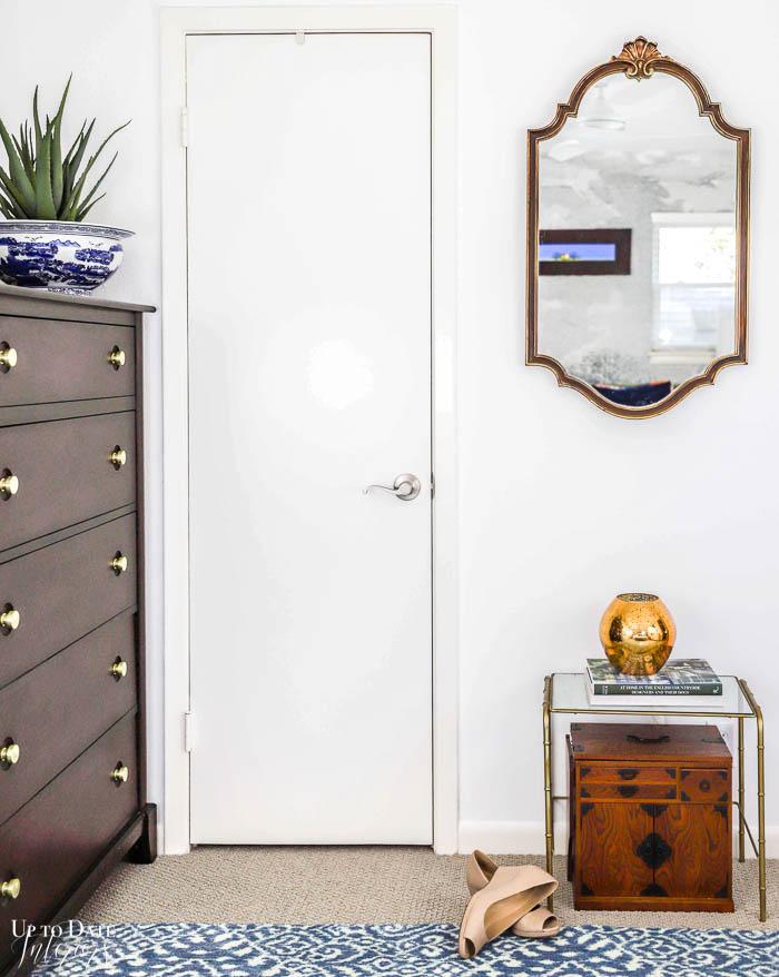 View of a bedroom closet door with bright white walls and a blue and white modern rug.  A gold estate sale mirror hangs above a glass and brass bamboo accent table with a Japanese writing desk underneath.  To the side is a tall wooden dresser with an aloe vera plant in a chinoiserie bowl. 
