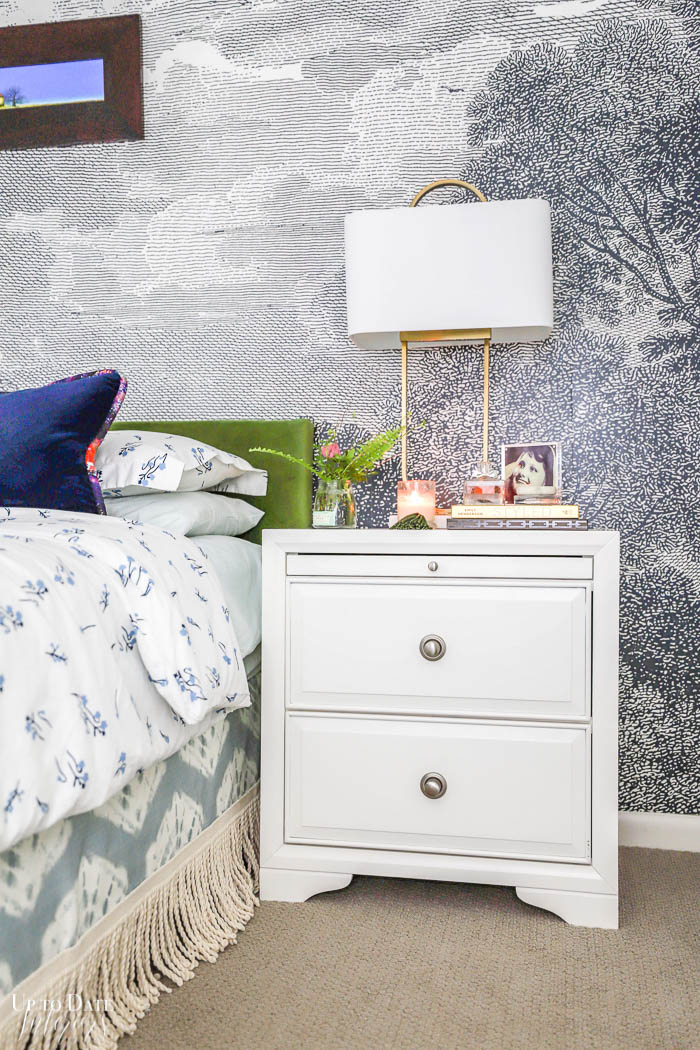 A modern white night stand is next to a bed with blue and white floral duvet, shibori and fringe bed skirt and a velvet green headboard.  A grey and white mural is on the wall. 