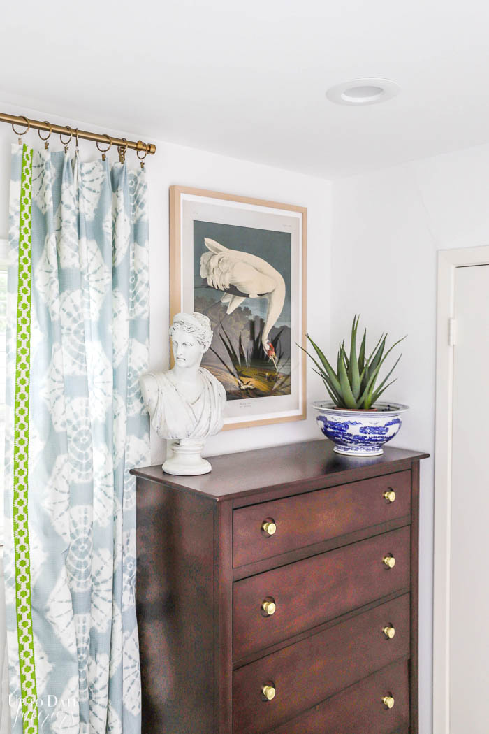 A tall dark dresser is updated with brass pulls and looks beautiful styled with a woman's bust, chinoiserie planter with aloe plant, and a vintage bird art print next to blue and white shibori curtains. 