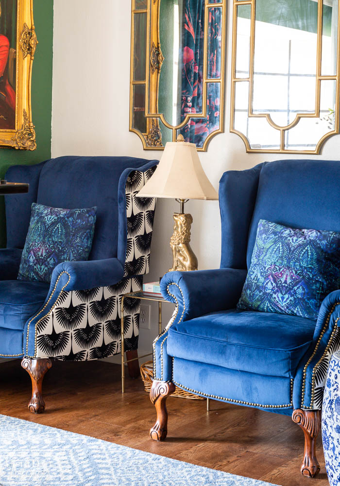A close up view of a pair of blue velvet wing back chairs with blue global throw pillows.  A small bamboo table with a brass lion lamp is in between and a pair of gold mirrors hang on the white wall. 